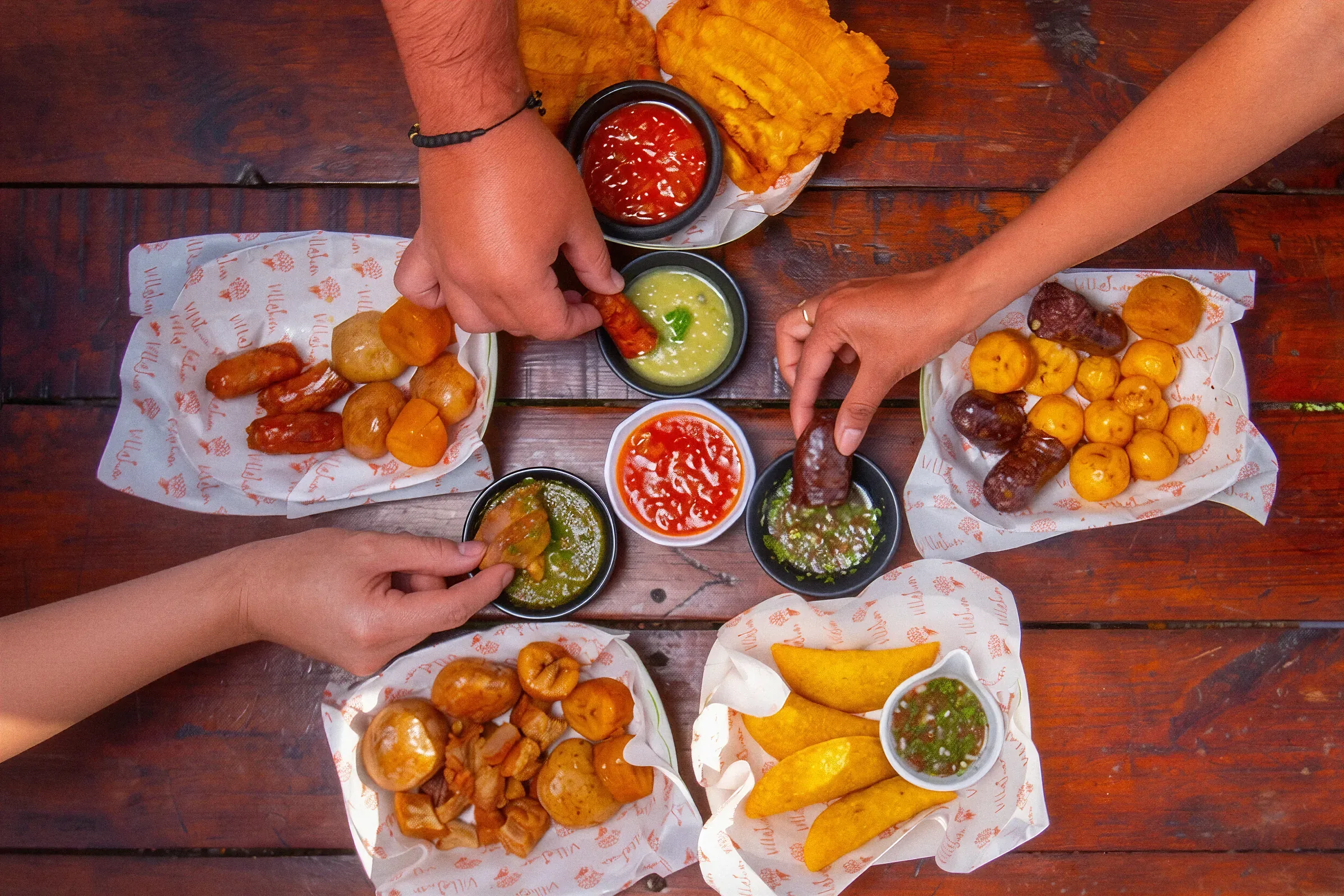 Manos compartiendo una picada de empanadas, patacones, chorizo y salsas en la mesa de madera de la Ecogranja Villa Juan